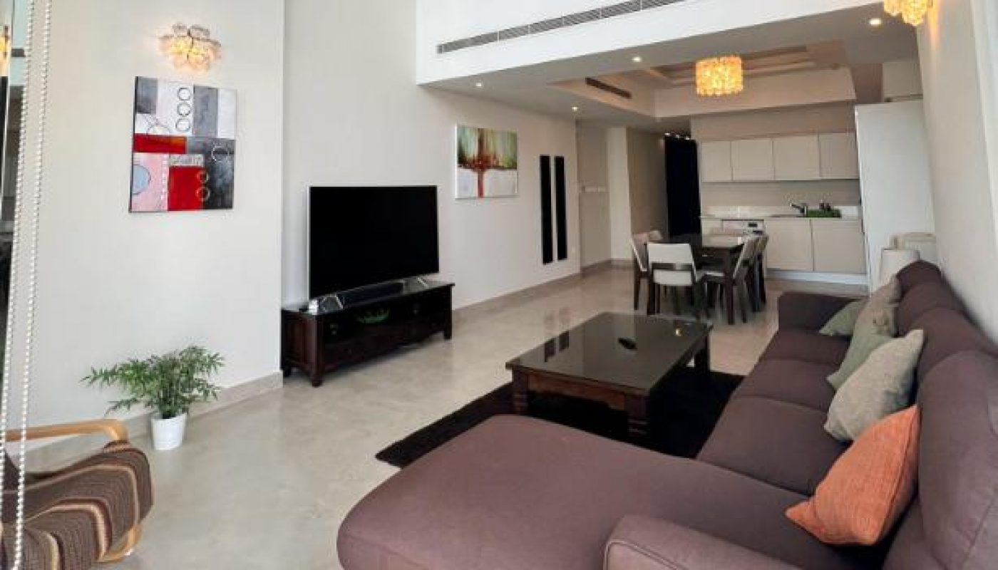Modern living room with high ceiling, brown sectional sofa, coffee table, wall-mounted TV, abstract art, potted plant, dining area, open kitchen, and large windows with natural light.