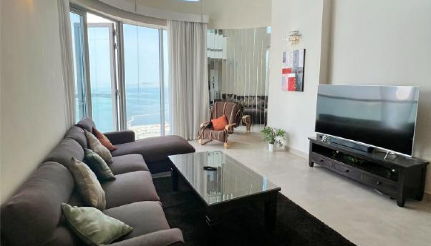 Modern living room with high ceilings, large windows, gray sofas, glass coffee table, flat-screen TV, and balcony view of the sea.