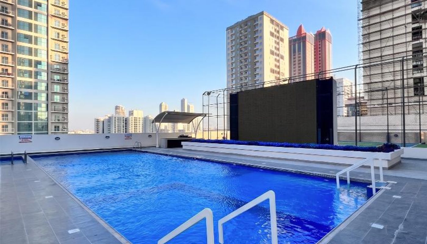 Rooftop swimming pool with clear blue water, surrounded by tile flooring and railings, with high-rise buildings visible in the background under a clear sky.
