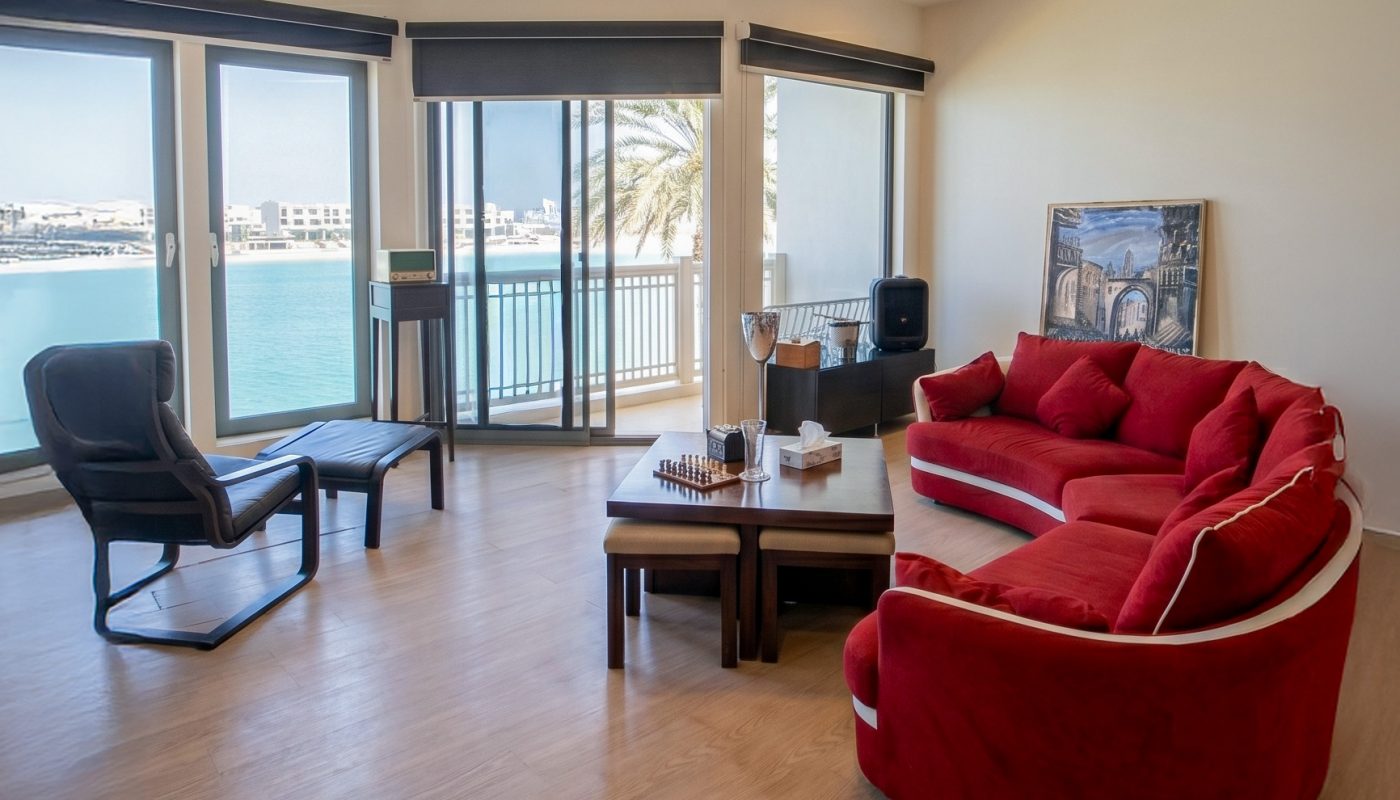 Modern living room with wood flooring, red sofa, black armchair, coffee table, large windows, and a view of water and palm trees outside.