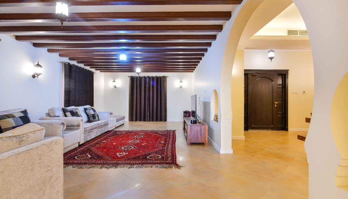 Spacious living room with beige sofas, a red patterned rug, wooden ceiling beams, arched doorway, and tiled floor, illuminated by multiple wall and ceiling lights.