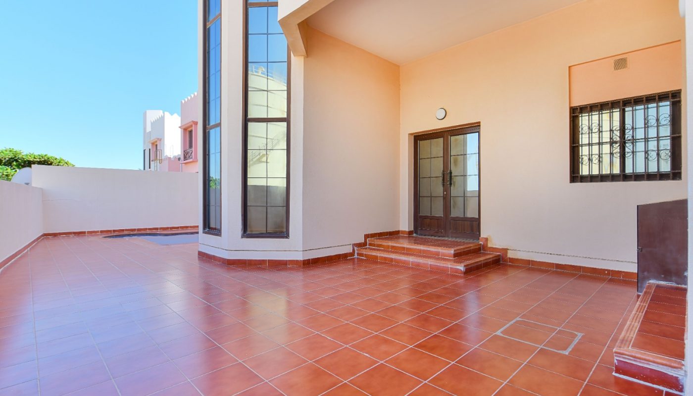 Spacious outdoor patio with red tile flooring, a covered entryway with stairs, large windows, and cream-colored walls under a clear blue sky.
