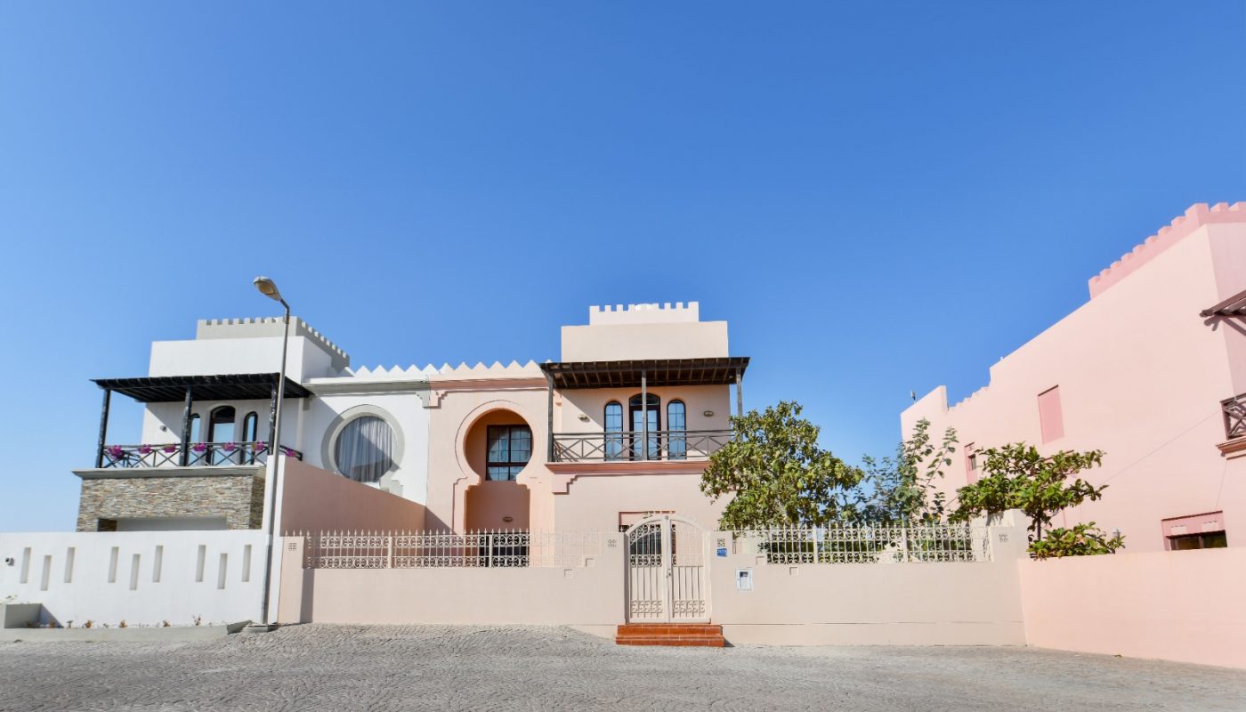 A modern, pastel-colored house with arched windows and a gated entrance sits under a clear blue sky on a cobblestone street.