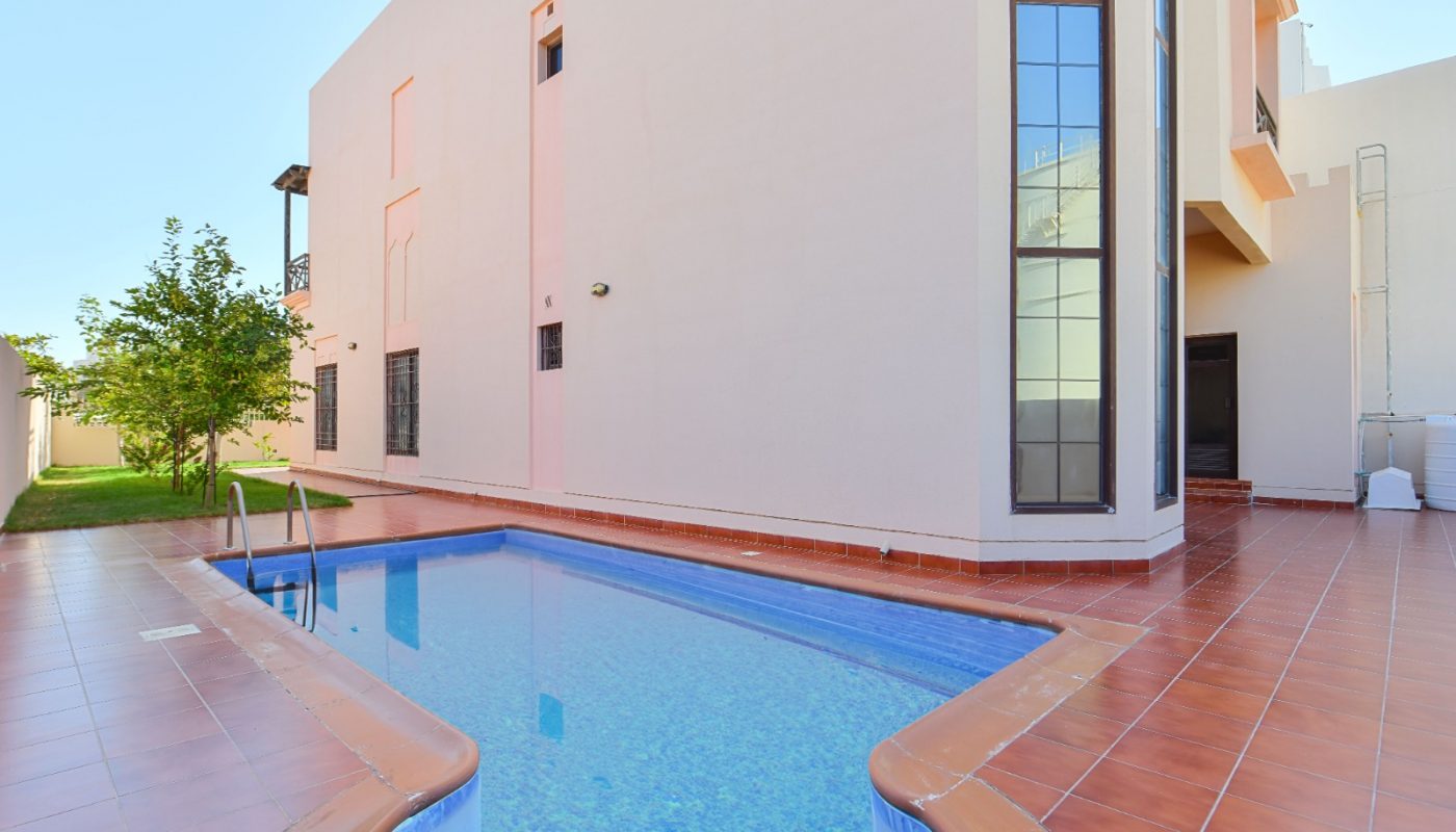 Rectangular outdoor swimming pool beside a modern beige building with large windows, surrounded by red tile flooring and a small grassy area with a tree.