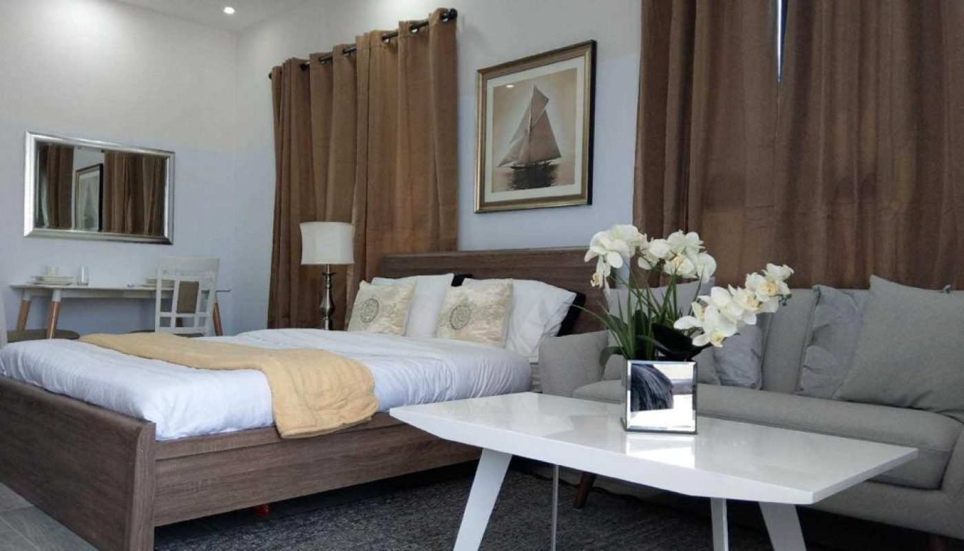 A modern bedroom with a double bed, beige curtains, a gray sofa, a white coffee table with white orchids, and a small desk with a chair in the corner.