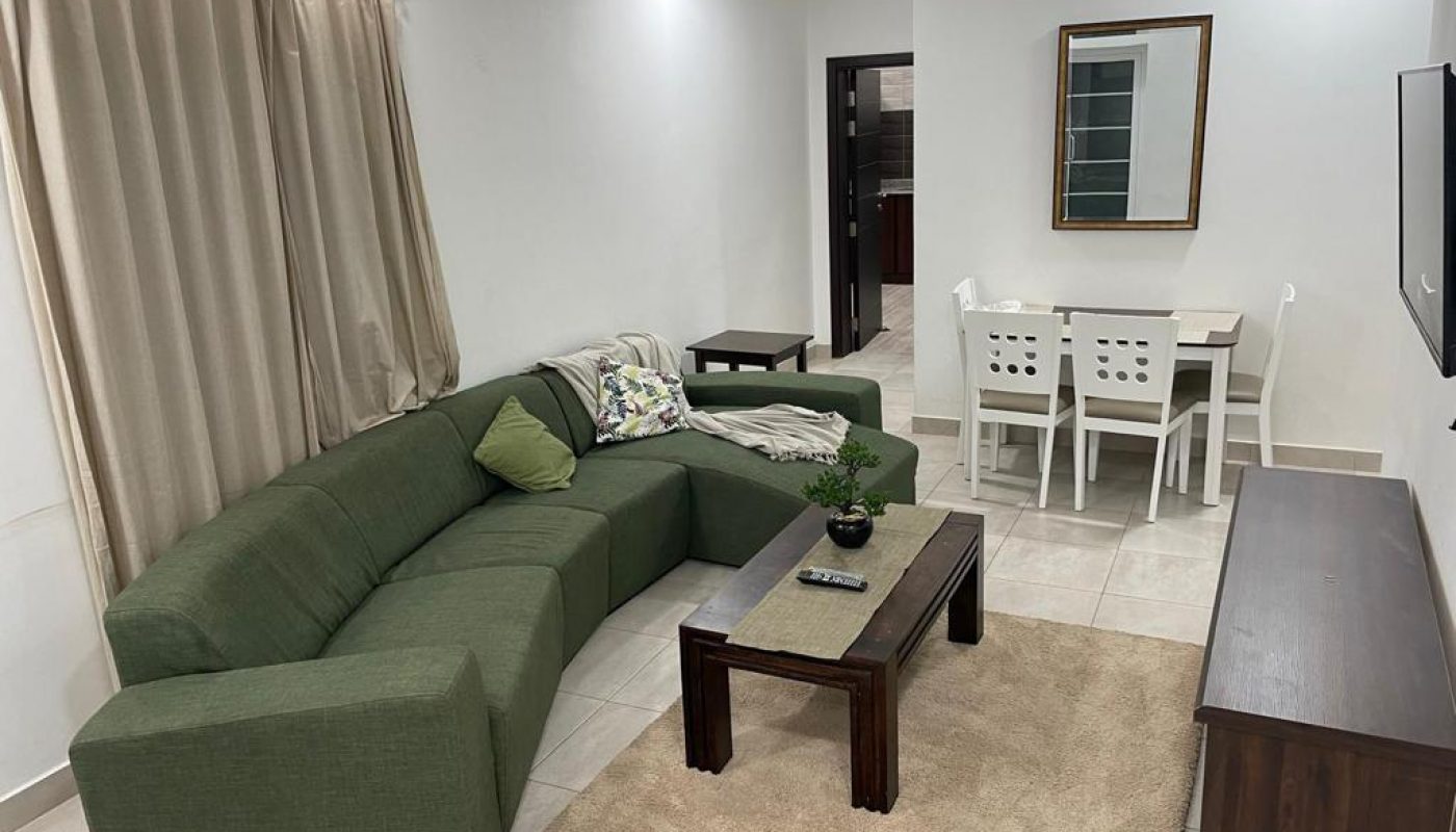 A living room with a green sectional sofa, coffee table, and TV, adjacent to a small dining area with a white table and four chairs. A beige curtain covers the window.