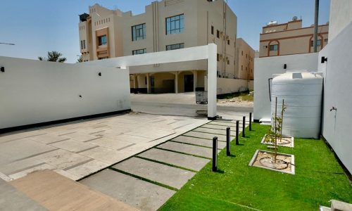 A clean, modern outdoor space with a paved area, green lawn, and two white buildings in the background under a clear blue sky.