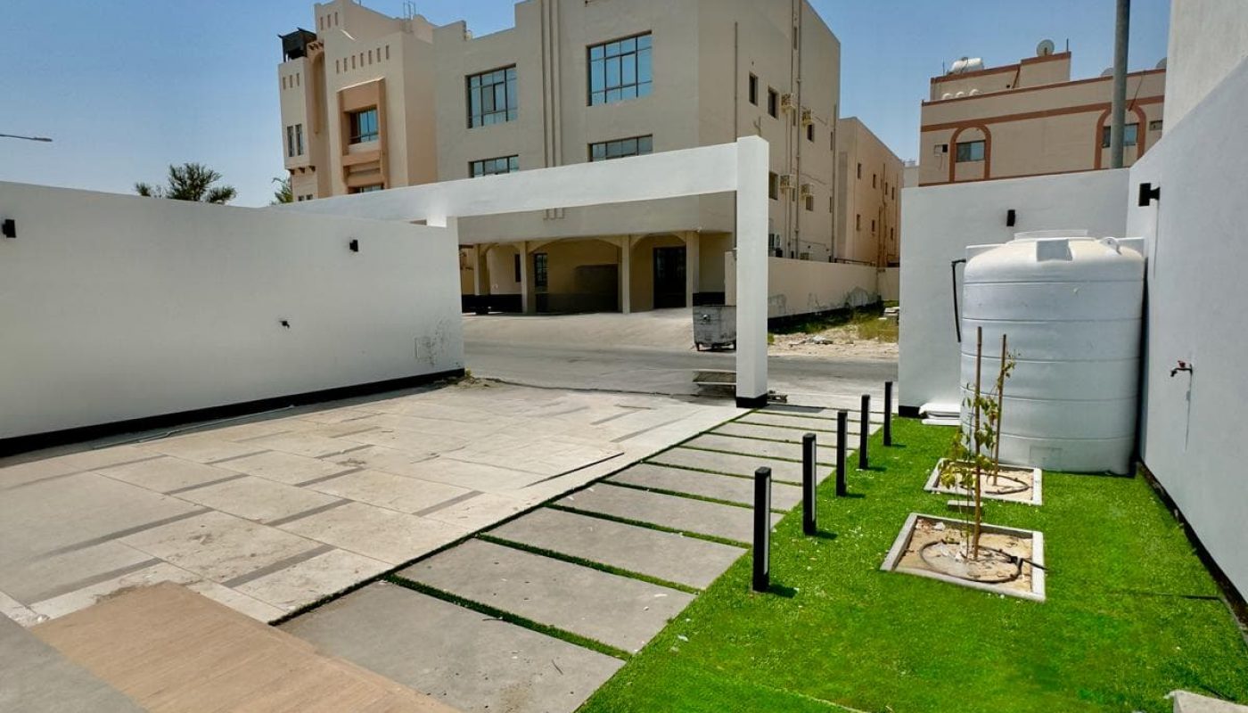 A clean, modern outdoor space with a paved area, green lawn, and two white buildings in the background under a clear blue sky.