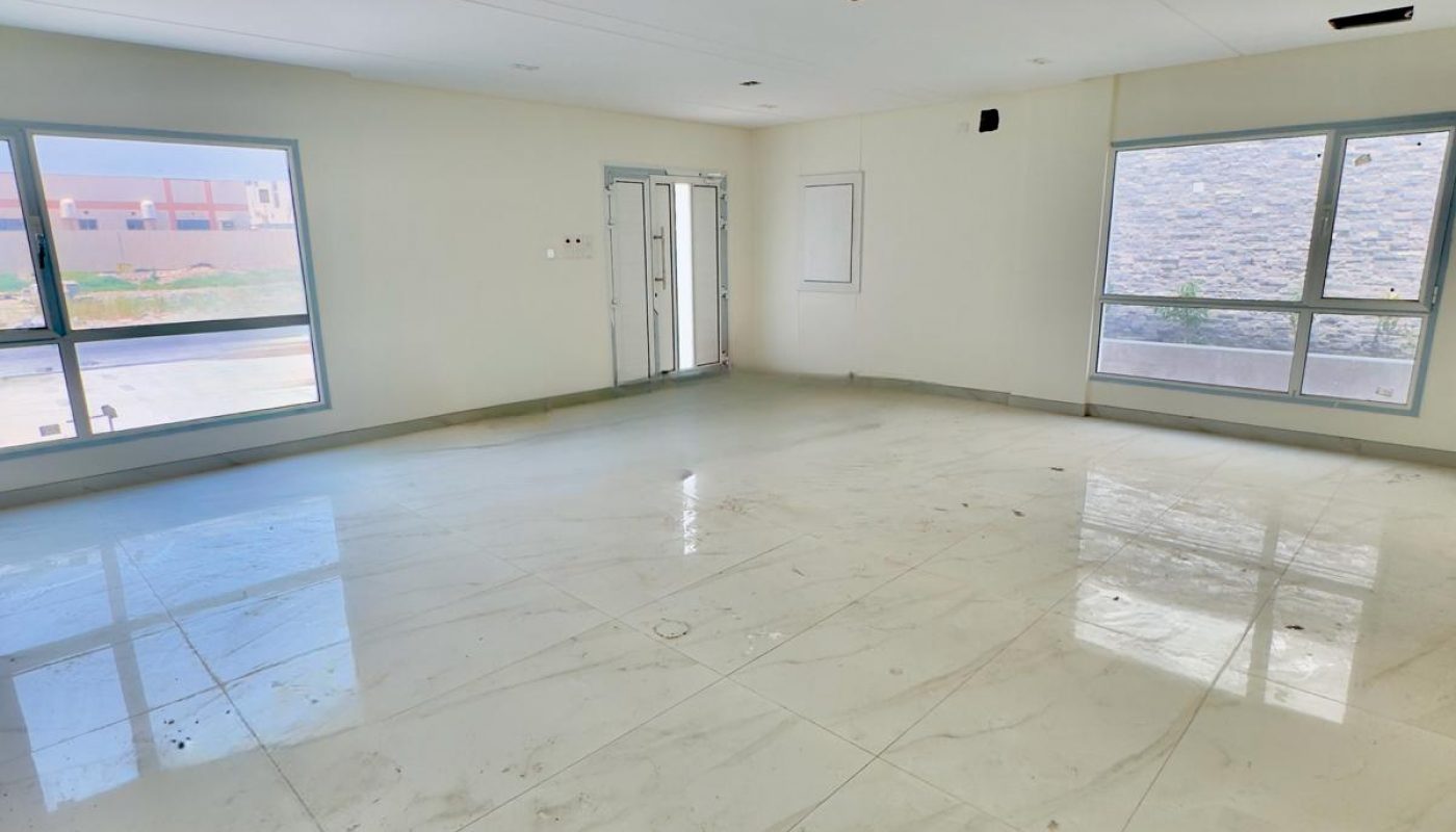 Spacious empty room with large tiled floor, white walls, and multiple windows allowing natural light. Door visible in the back corner.