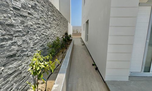 Narrow outdoor pathway with a stone wall on the left and a white building on the right. Small plants line the edge of the path under a clear blue sky.