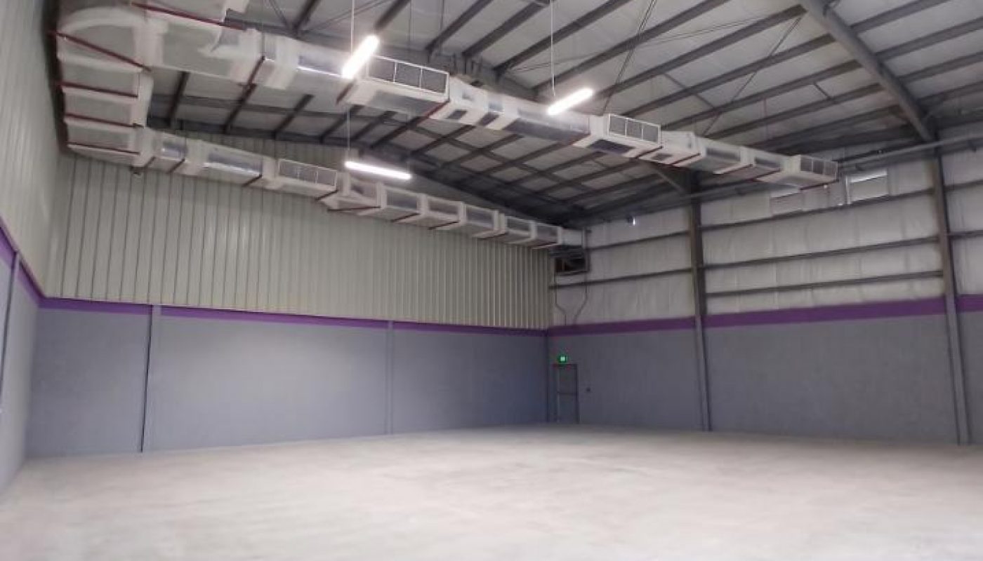Empty warehouse with high metal ceiling, exposed ductwork, concrete floor, and gray walls with a purple stripe. Fluorescent lights hang from the ceiling. A closed door is visible in the back wall.
