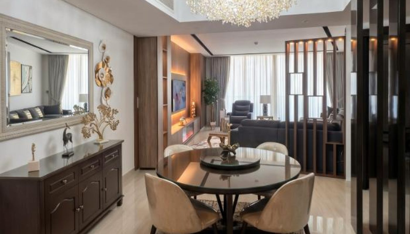 Modern dining area with a round table and four chairs, chandelier overhead, sideboard with decor, and open view into a living room with large windows.