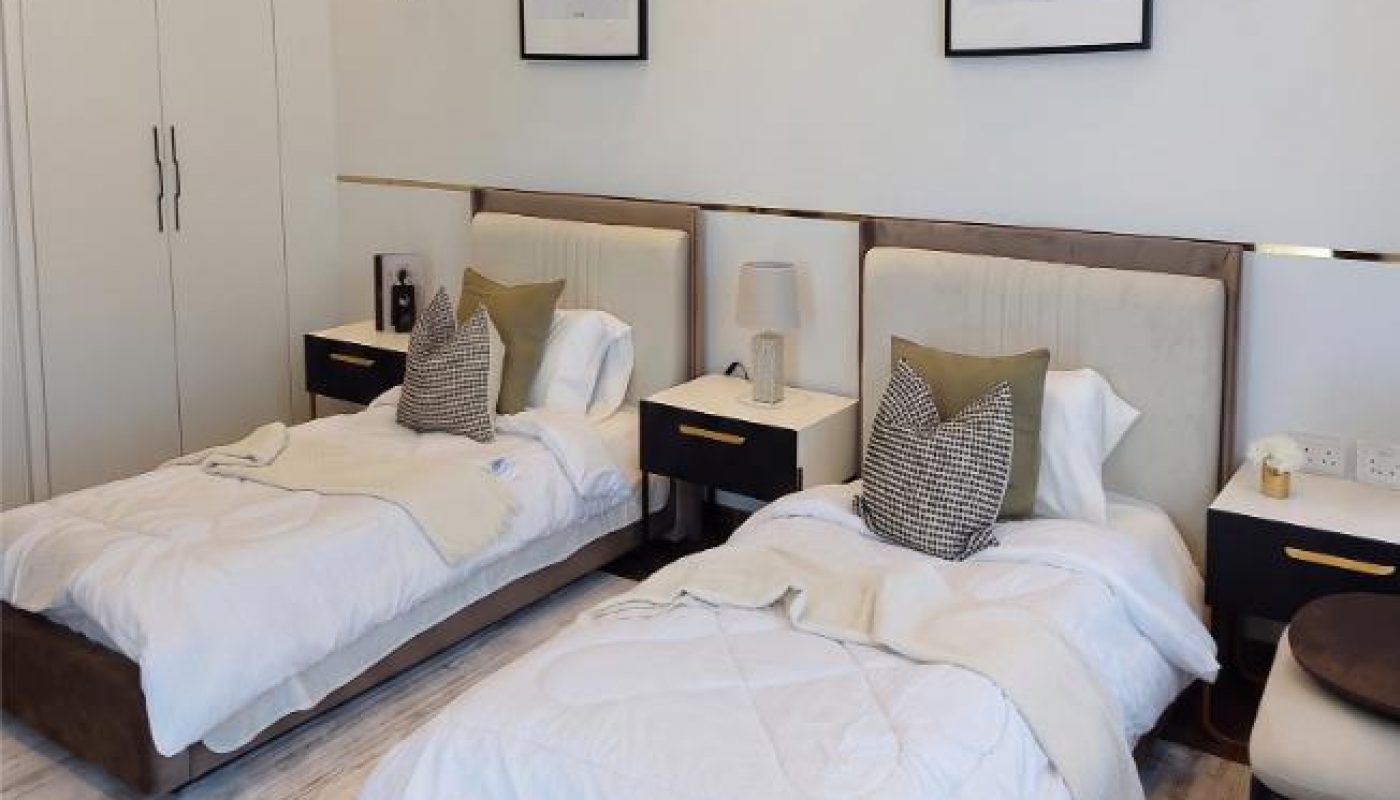 A modern bedroom with two single beds, matching bedding, dark nightstands, abstract art on the wall, and a large white wardrobe.