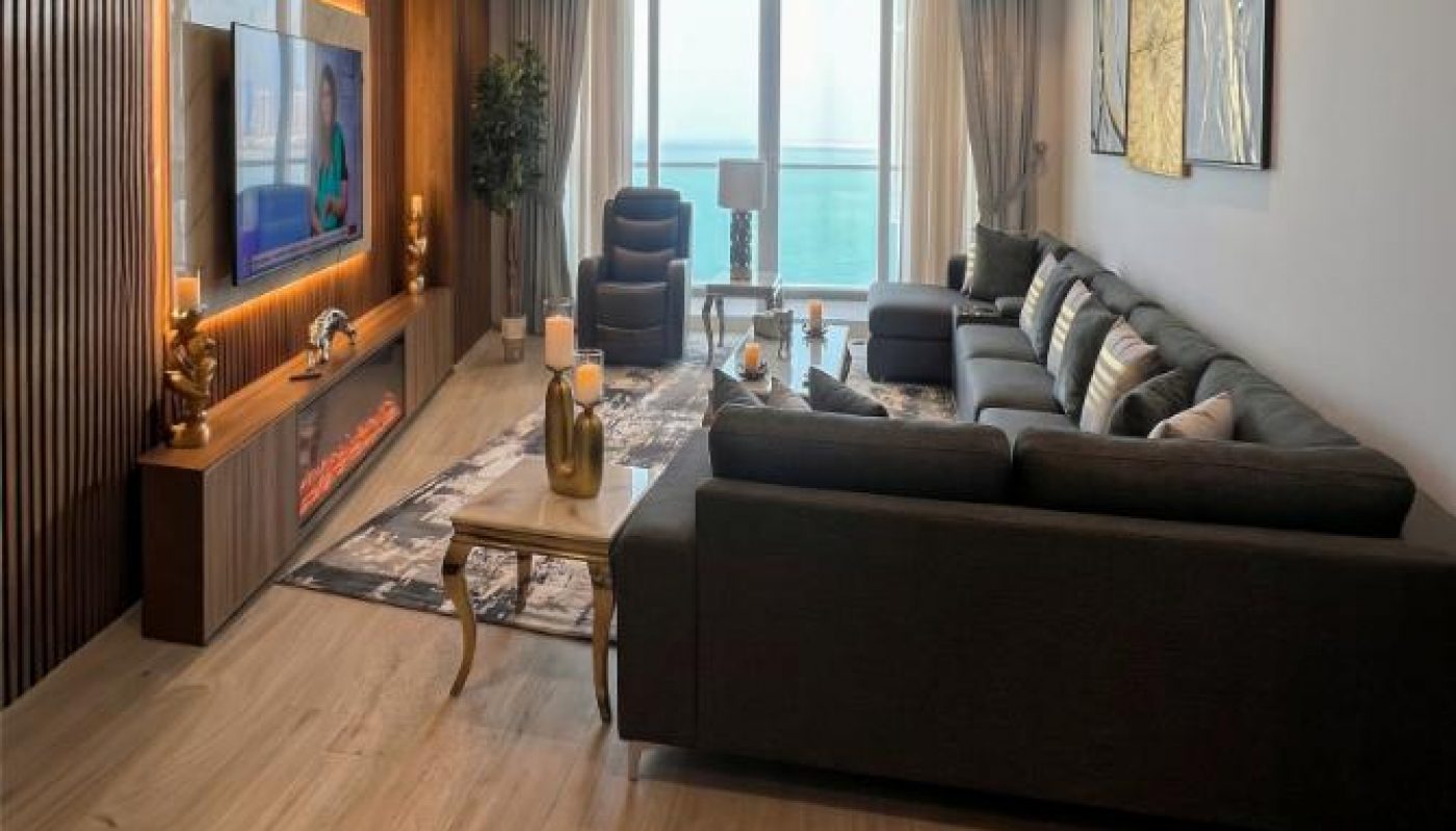 Modern living room with a large sectional sofa, wooden accent wall with mounted TV, coffee table, artworks, and floor-to-ceiling windows overlooking a balcony with an ocean view.