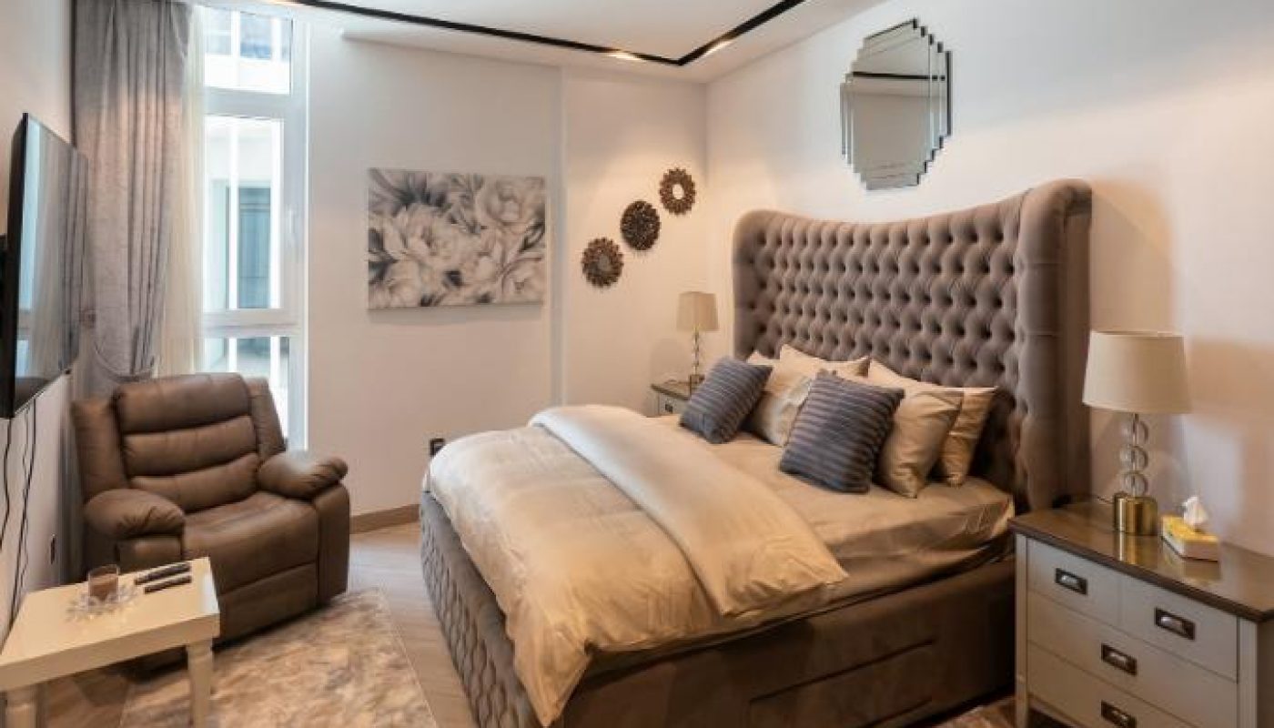 Modern bedroom with a tufted bed, armchair, side tables with lamps, wall art, and two rugs on a wooden floor under recessed ceiling lights.
