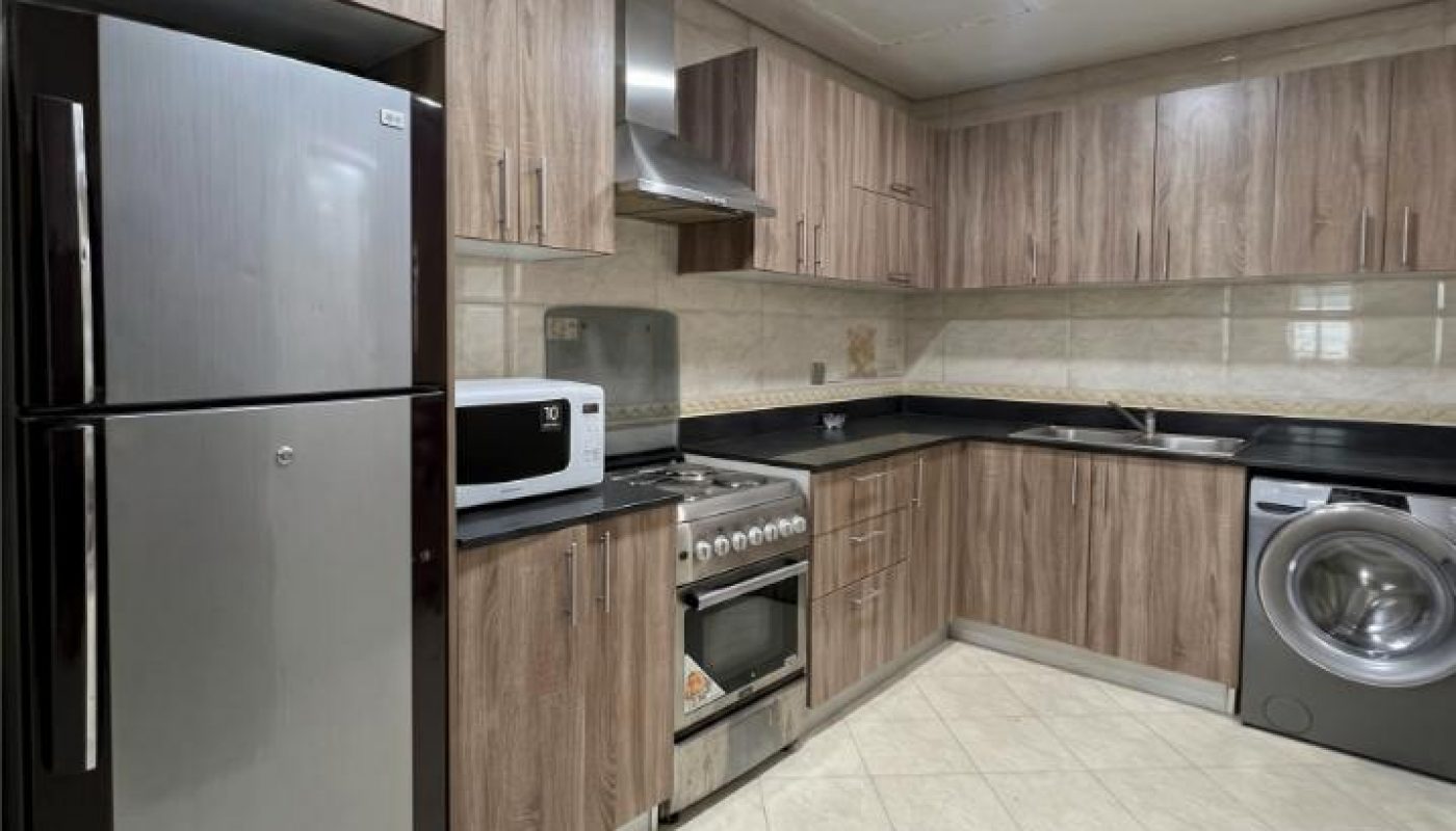 Modern kitchen with wood cabinets, stainless steel refrigerator, oven, microwave, washing machine, and tiled floor under a ceiling light.