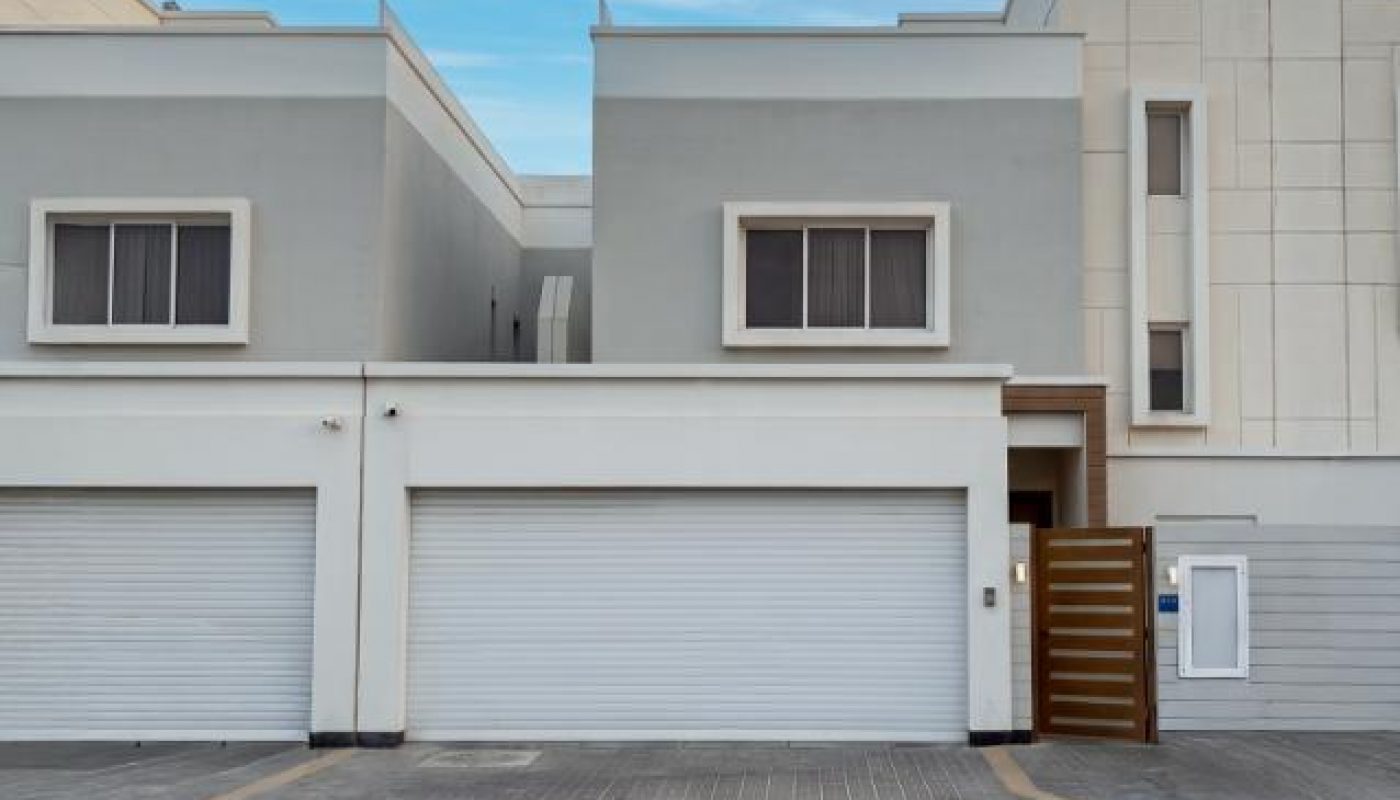 Modern two-story townhouse with grey and white exterior, three closed garage doors, and a small wooden gate, set against a clear blue sky.