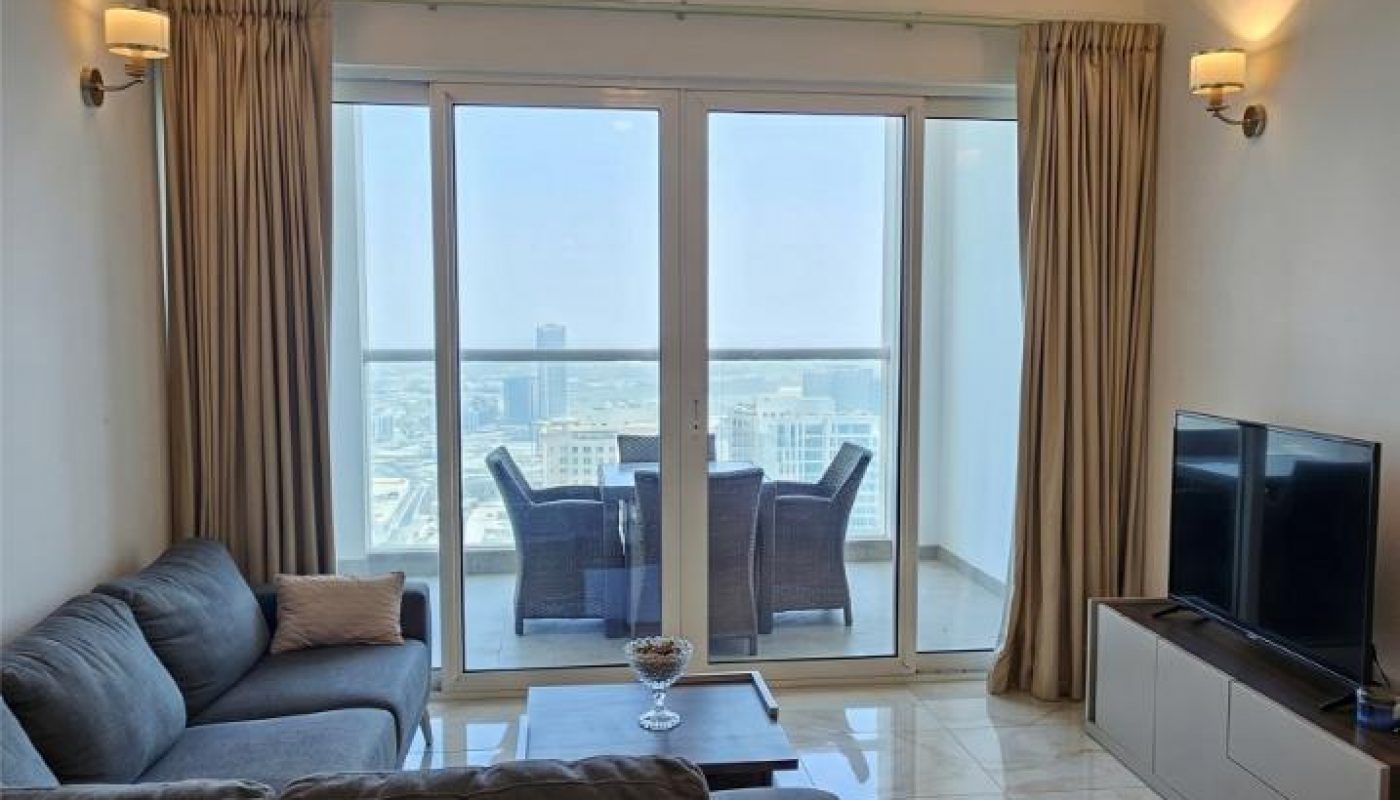Modern living room with a dark sectional sofa, glass coffee table, TV, and floor-to-ceiling windows opening to a balcony with city views and an outdoor dining set.