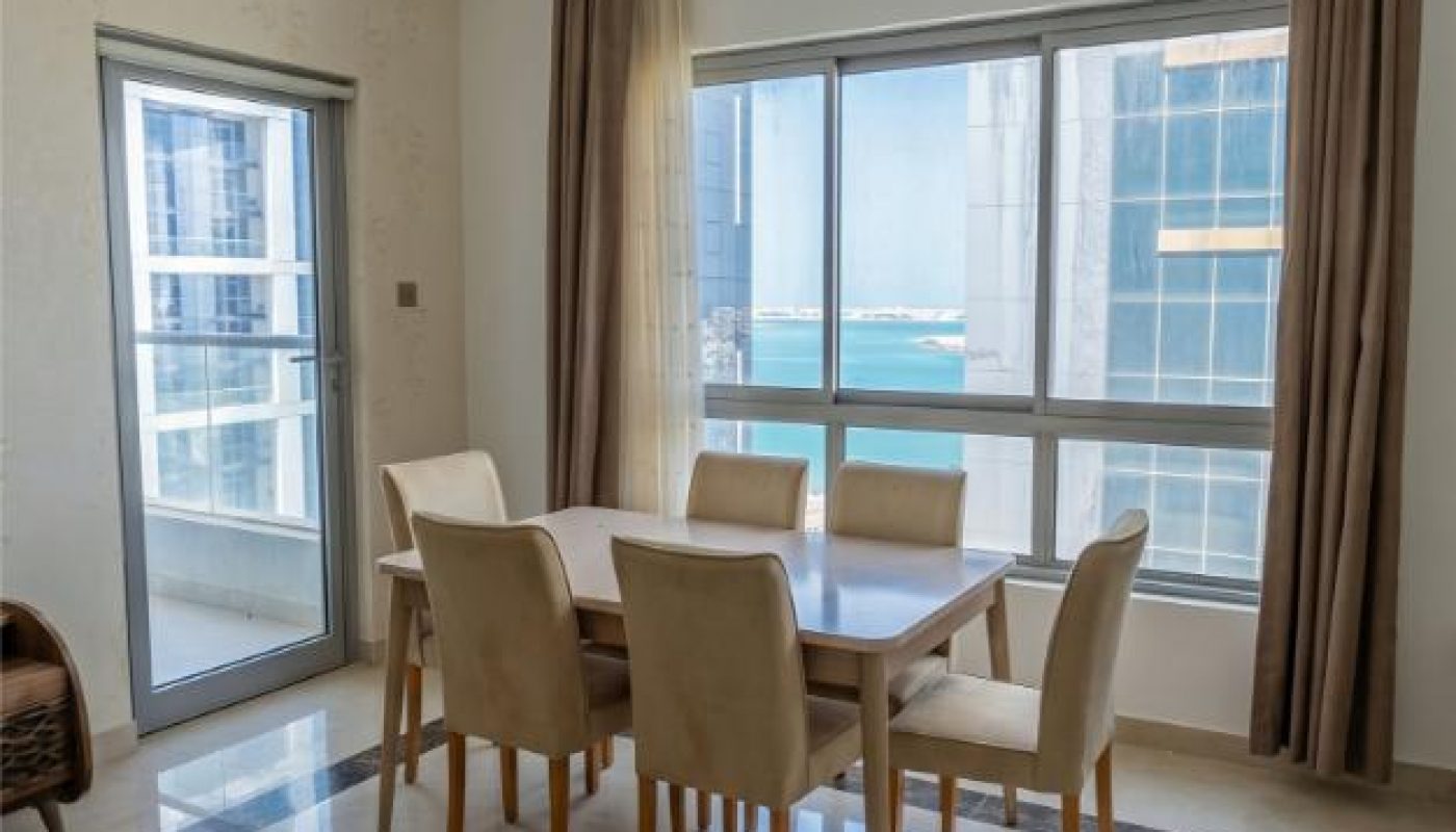 A modern dining room with a wooden table, six beige chairs, floor tiles with a dark border, large windows, and beige curtains.