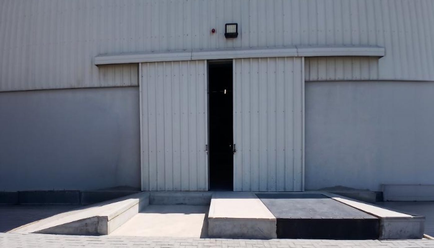 A large white industrial warehouse with a closed sliding door, loading docks, and a paved area in front, under a clear blue sky.