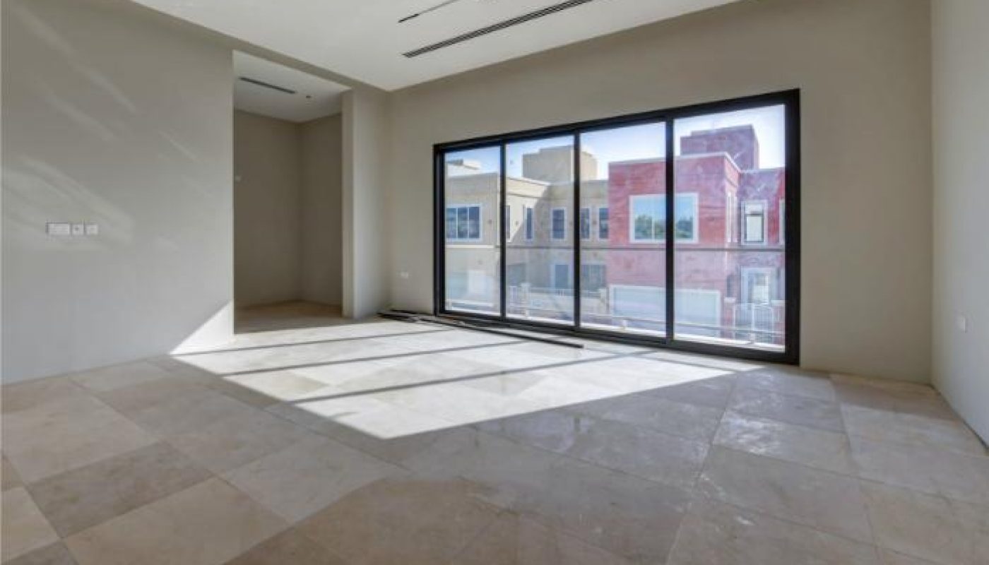 Empty room with large beige floor tiles, off-white walls, ceiling lights, and a full-length window overlooking buildings outside.