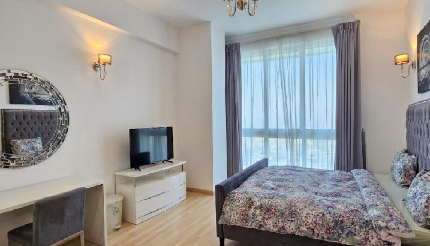 A modern bedroom with wood flooring, a bed with floral bedding, a TV on a white dresser, a mirror above a desk and chair, and large windows with sheer curtains.