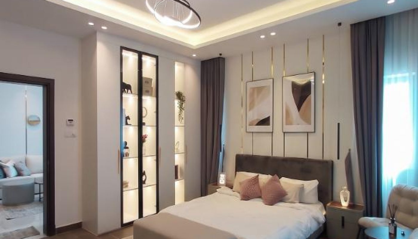 Modern bedroom with a double bed, accent chairs, wall art, floor-to-ceiling curtains, and built-in shelving, featuring neutral colors and soft lighting.