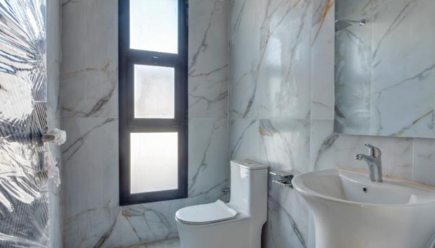 A modern bathroom with marble walls and floor, a wall-mounted sink, a toilet, a large mirror, a window, and a showerhead.