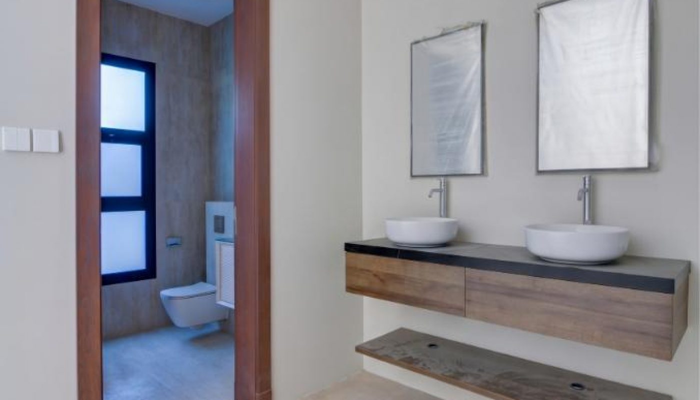 Modern bathroom with beige tile flooring, a double sink vanity with two mirrors, and a separate toilet area visible through a wooden-framed doorway.