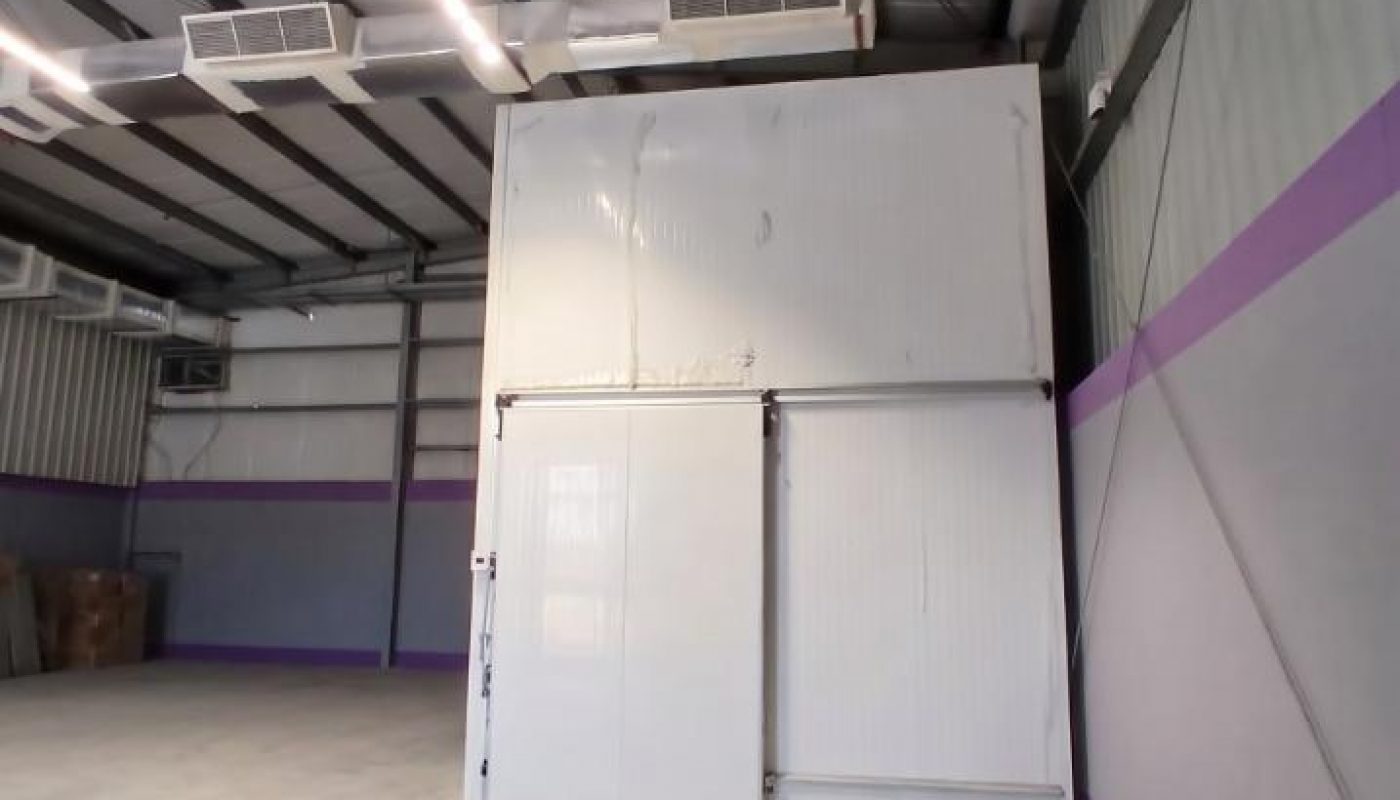 Large empty warehouse interior with metal ceiling, exposed ducts, and a white walk-in cooler or storage unit against a wall with purple stripes.