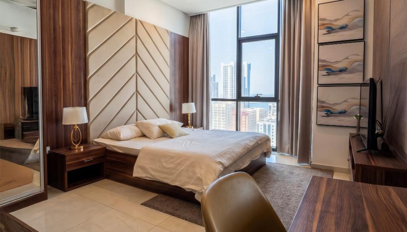 Modern bedroom with a large bed, wooden furniture, desk and chair, city view through tall windows, and decorative wall art.