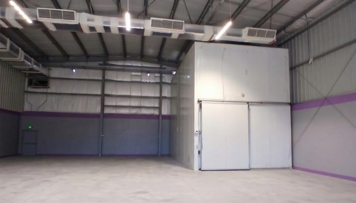 Interior of an empty industrial warehouse with concrete floors, metal walls, exposed beams, and a large white storage unit against the back wall.