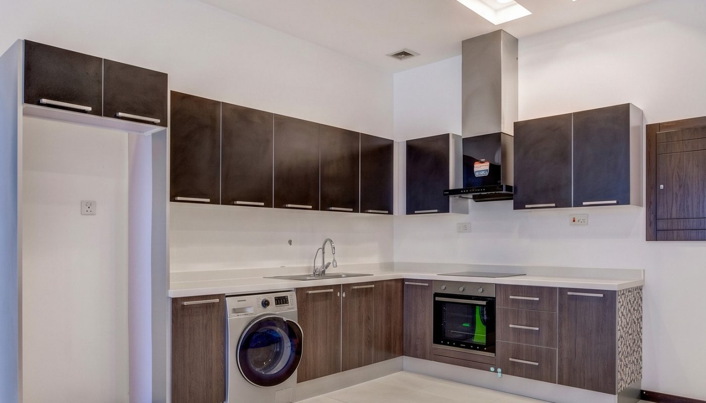 Modern kitchen with dark wood cabinets, white countertops, built-in oven, stovetop, washing machine, and under-cabinet sink on light tiled floor with recessed ceiling lights.