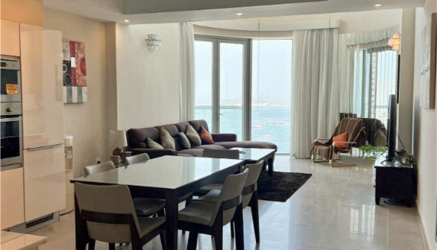 Modern open-plan living and dining area with large windows, a dining table, sofa, and a view of the sea in the background.