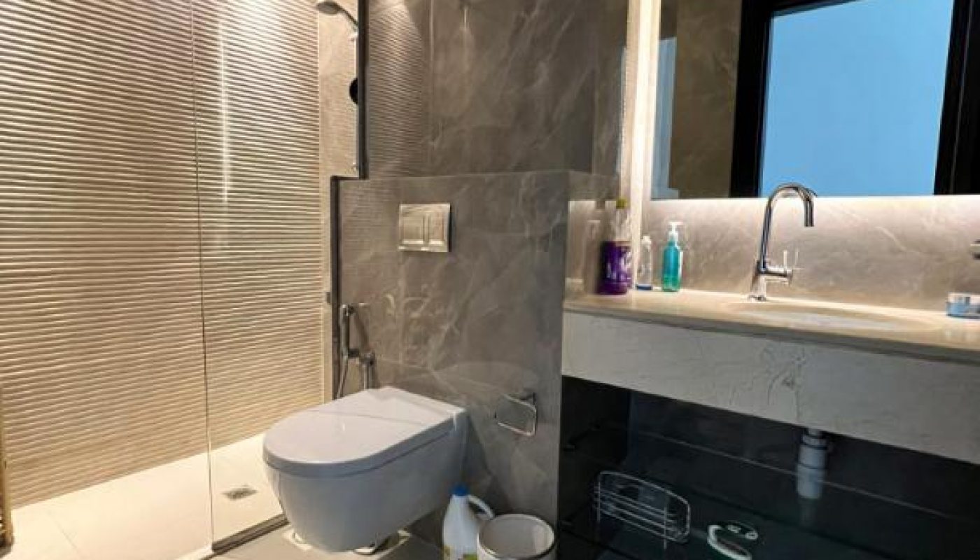 Modern bathroom with gray tiles, wall-mounted toilet, glass shower enclosure, large mirror, sink with toiletries, and cleaning supplies near a small trash bin.