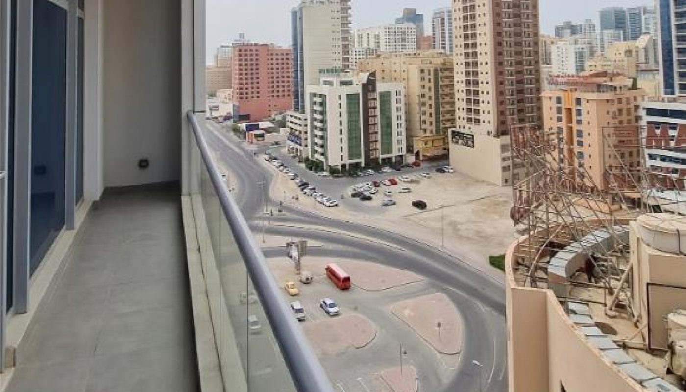 View from a high-rise balcony overlooking a city street with mid-rise buildings, parked and moving cars, and cloudy sky above.