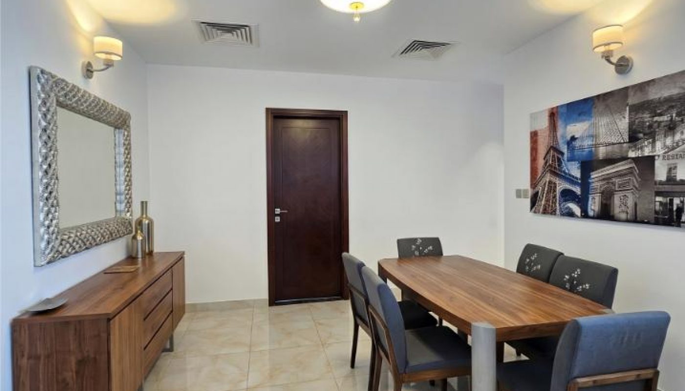A modern dining room with a wooden table, six chairs, a sideboard, a large mirror, wall art, and tiled floor. A closed wooden door and overhead lighting are visible.