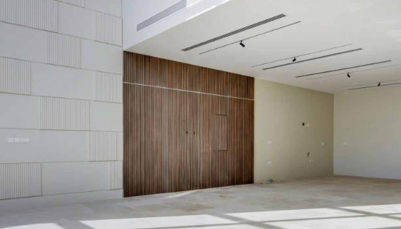 Large, empty modern room with high ceiling, tiled floor, wood accent wall, and sunlight streaming through large windows, casting shadows on the floor.