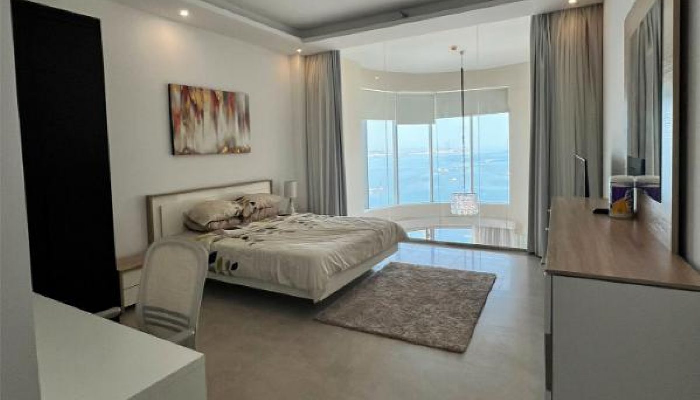 Modern bedroom with a bed, desk, chair, rug, dresser, wall art, and large windows offering an ocean view.