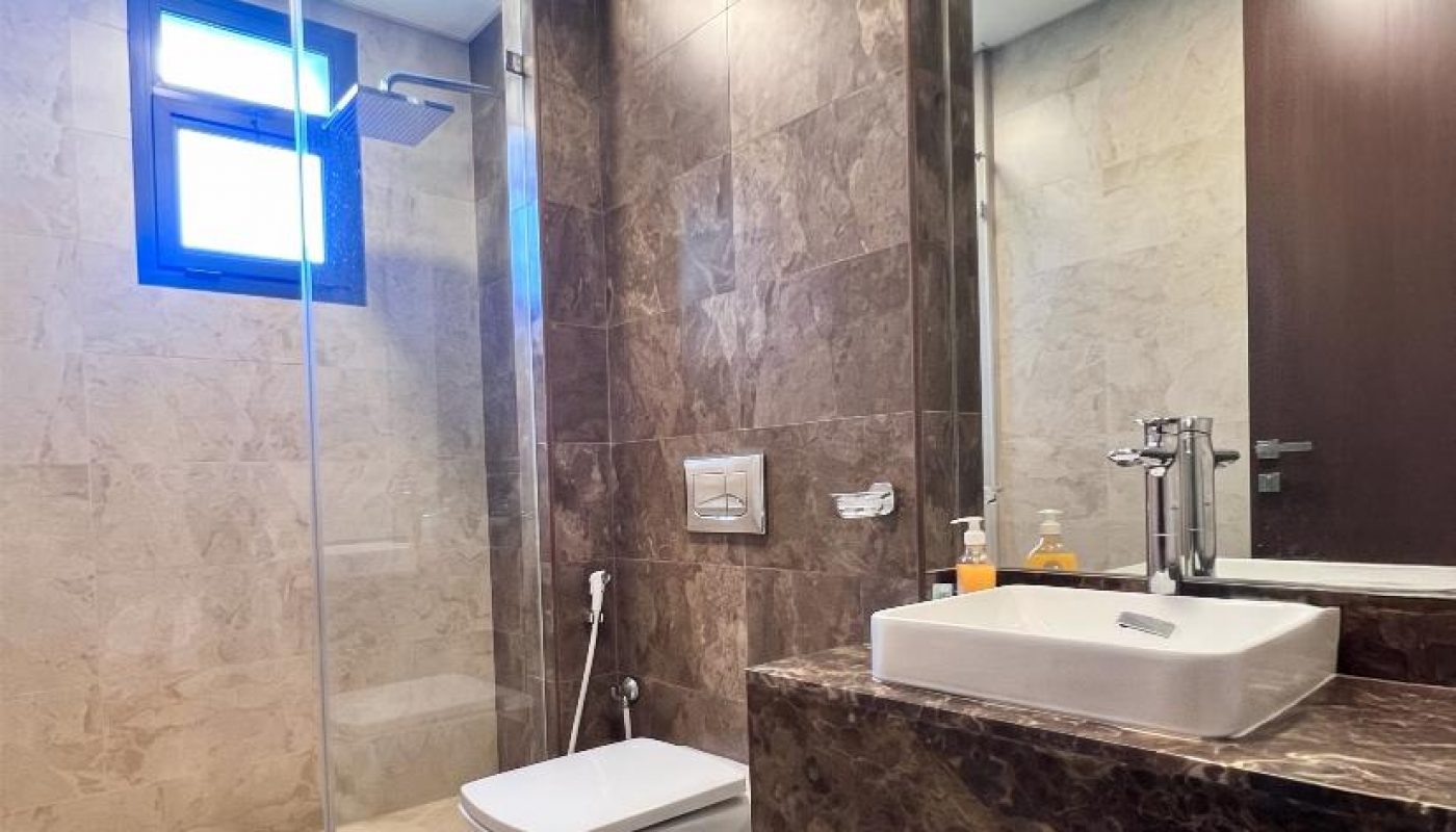Modern bathroom with marble walls, a wall-mounted toilet, a glass shower enclosure, a sink with a mirror, soap dispensers, a trash bin, and two white and black rugs.