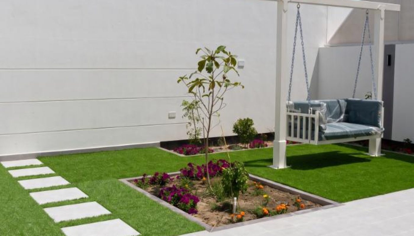 A modern backyard with artificial grass, stepping stones, a small garden bed with flowers, and a white porch swing with cushions.