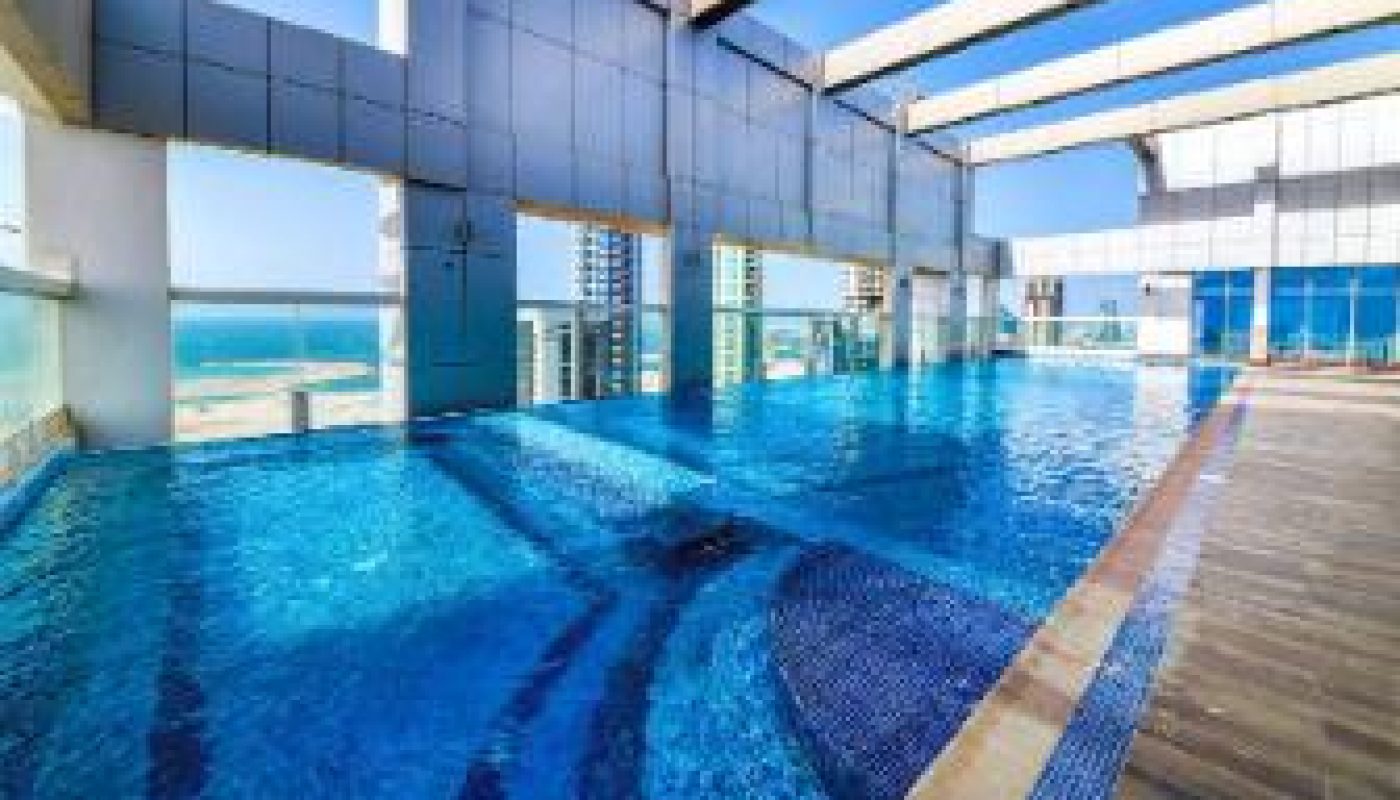 Rooftop swimming pool with clear blue water, surrounded by modern architecture and glass panels, overlooking a coastal cityscape under a clear sky.