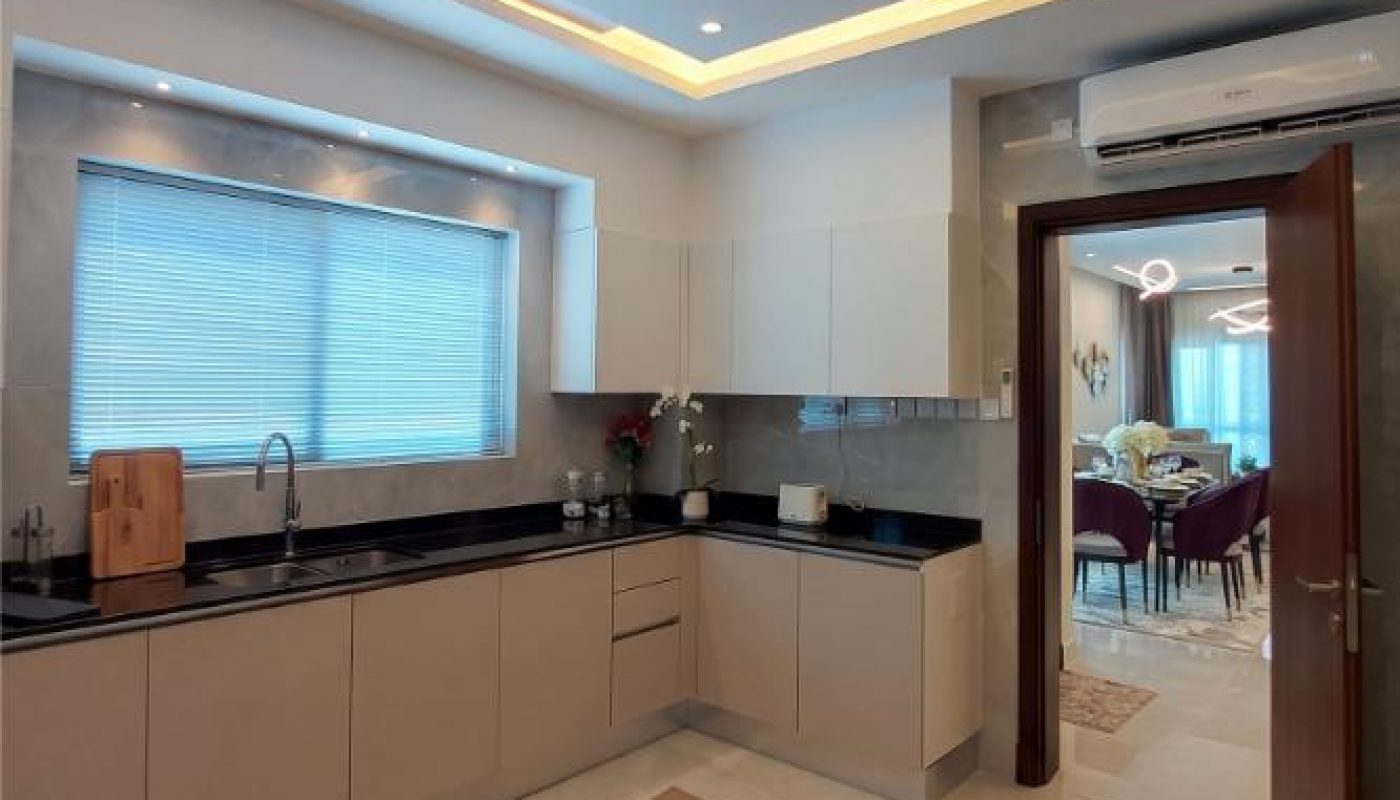 Modern kitchen with beige cabinets, black countertops, under-cabinet lighting, a large window with blinds, and an open door leading to a dining area.