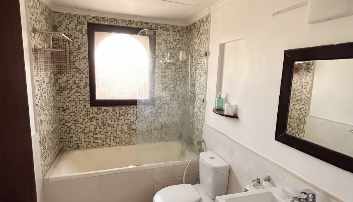 Modern bathroom with tiled bathtub, glass partition, toilet, sink, mirror, and window letting in natural light.