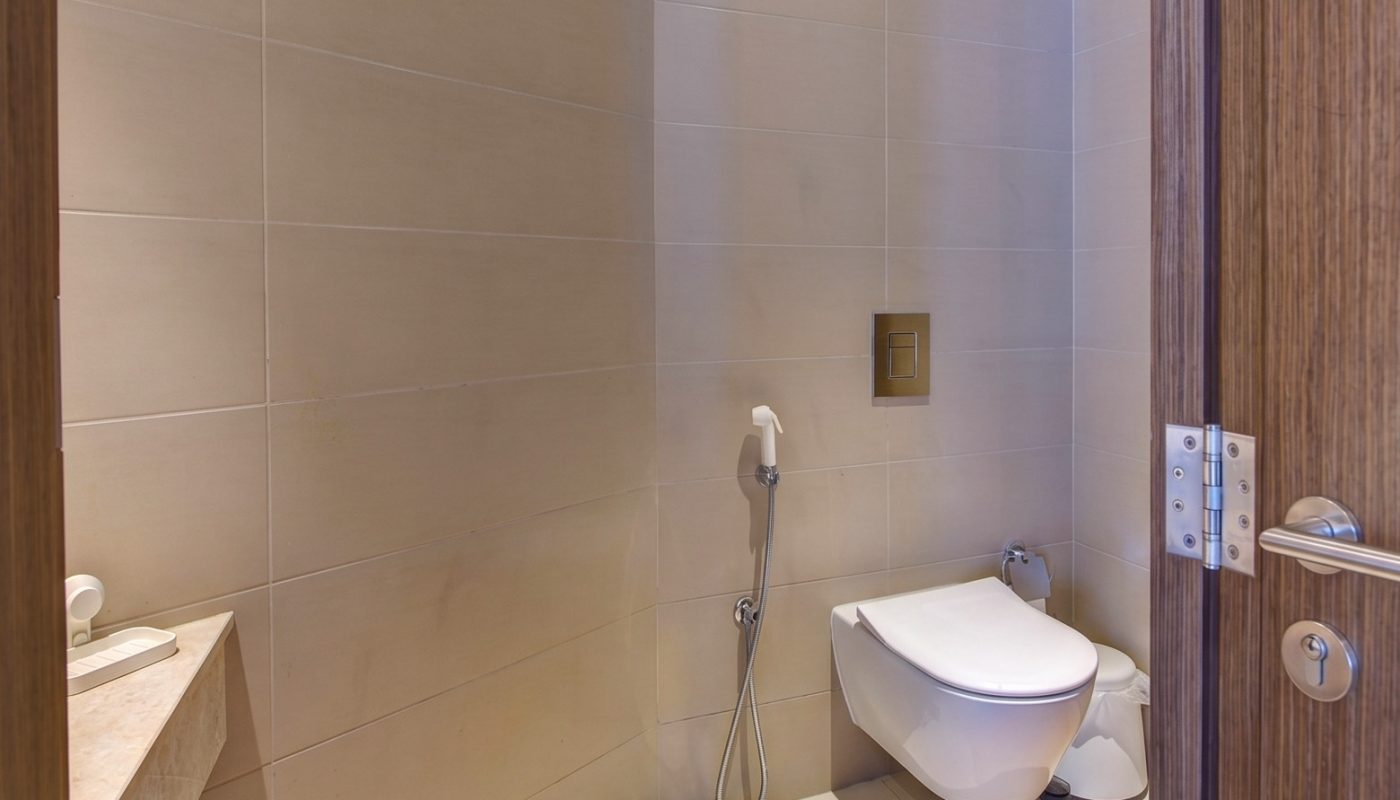 Modern bathroom with beige tiled walls, a wall-mounted toilet, handheld bidet spray, and a sink partially visible on the left.