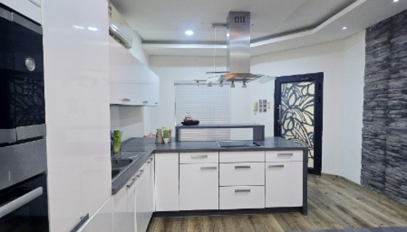 Modern kitchen with white cabinets, built-in ovens, an island with stove and range hood, wood-style flooring, and a decorative black door.
