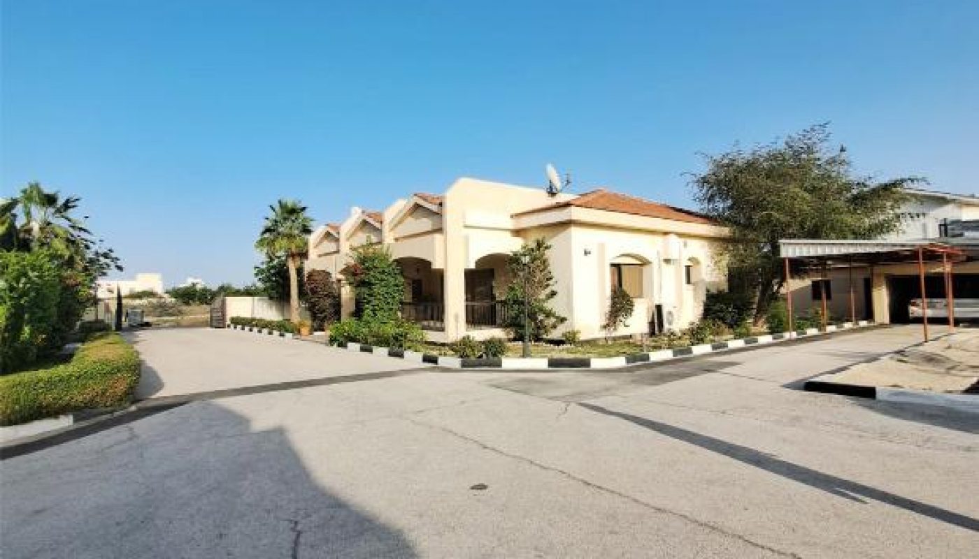 A large beige house with a red-tiled roof sits on a corner lot with a driveway, surrounded by greenery under a clear blue sky.