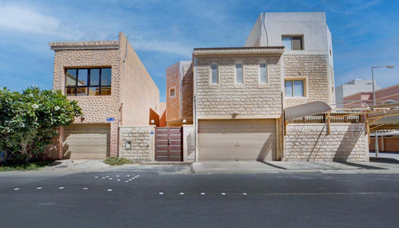 Two modern houses with stone facades and garages stand side by side under a blue sky, with a paved road in the foreground.
