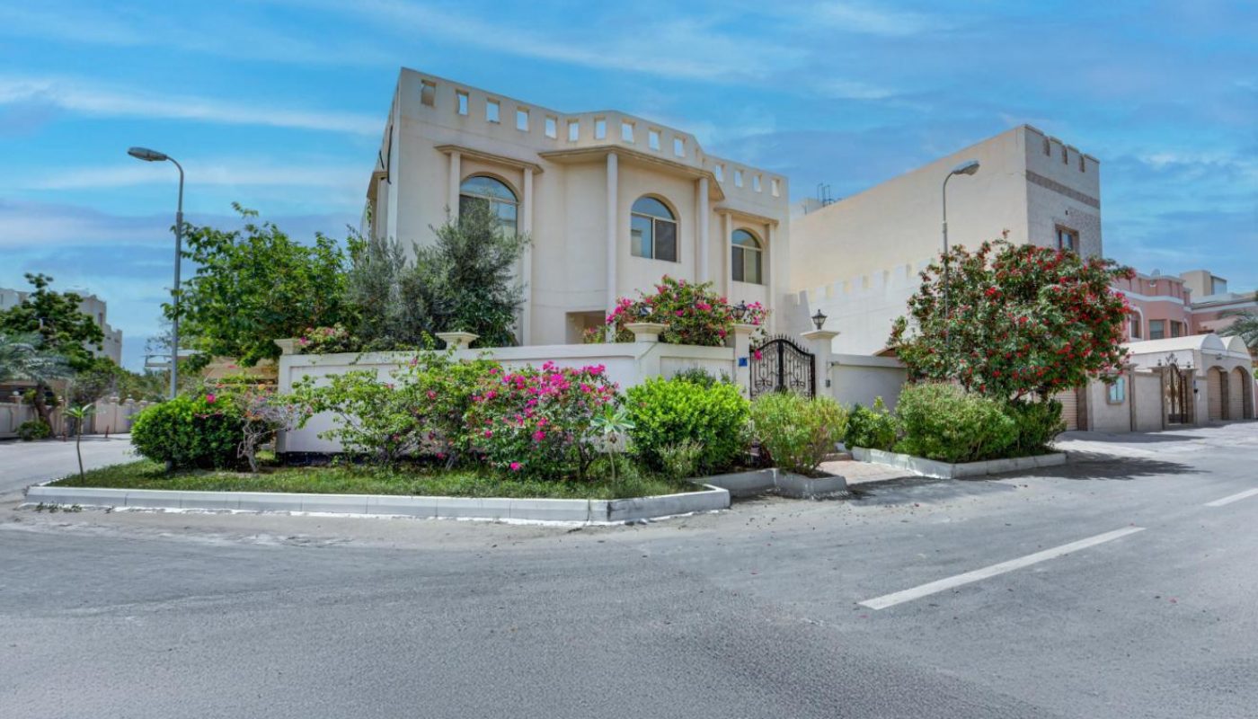 A beige two-story villa with arched windows and a landscaped garden sits on a corner lot under a partly cloudy blue sky.