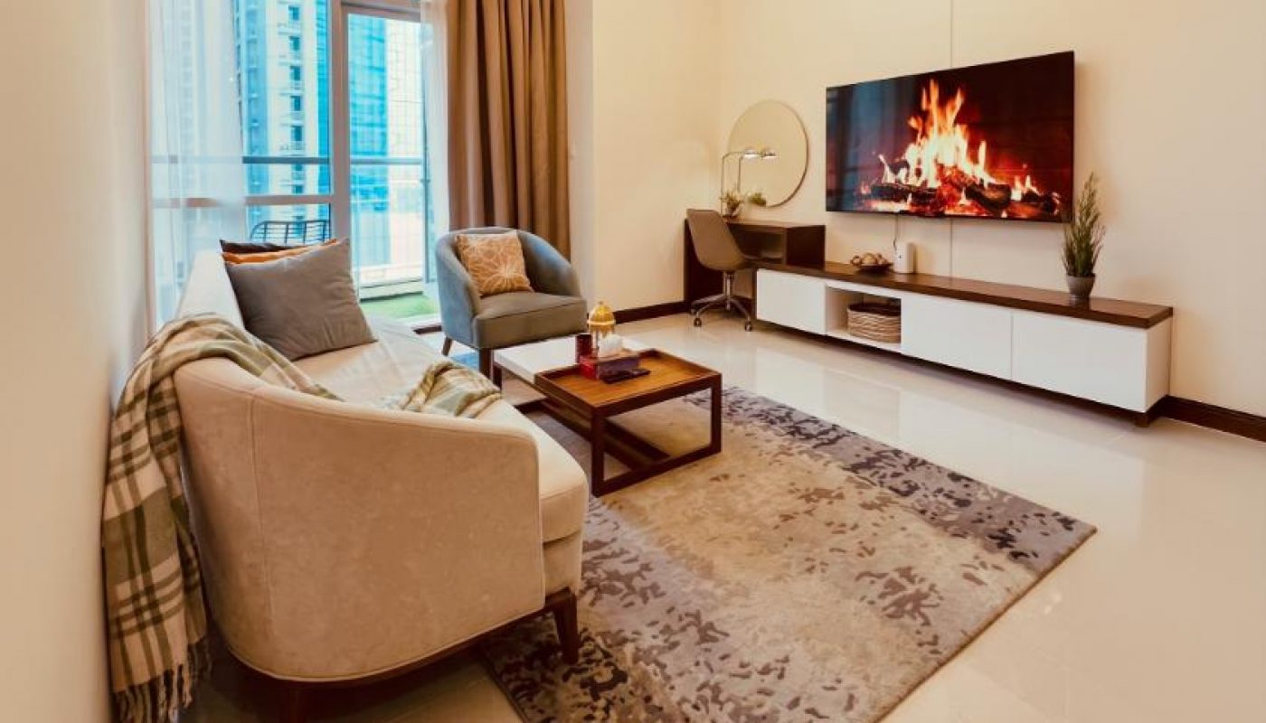 Modern living room with a sofa, armchair, coffee table, rug, TV displaying a fireplace, side table, large window, and city view.